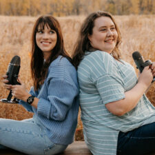 Two caucasian women wearing blue with brown hair sitting back to back on a bench holding microphones in front of a brown grassy field.